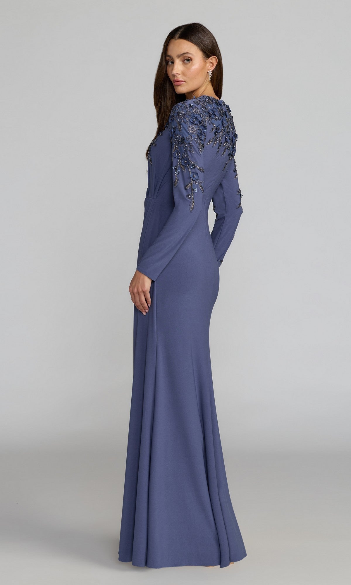 A woman stands and looks over her shoulder, wearing the Mac Duggal 13246 long formal dress—a floor-length, long-sleeved blue gown adorned with floral embellishments on the shoulders and back.